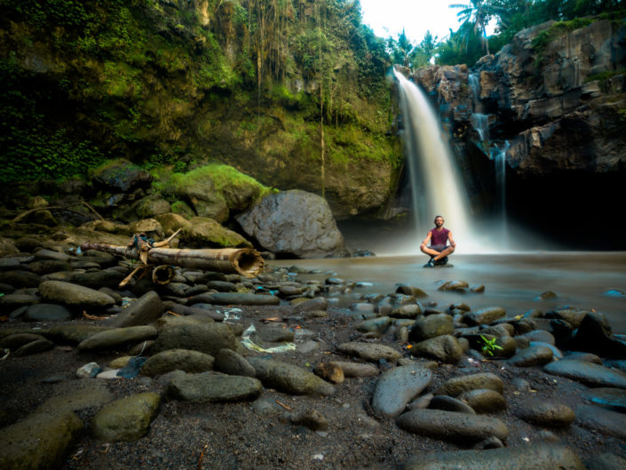 Man sits cross legged on rocks beneath a flowing waterfall